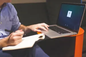 A person writing on a laptop with a notebook